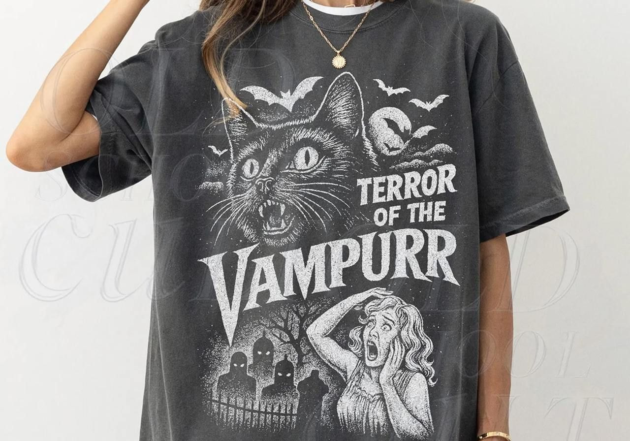 A woman in a gray t-shirt with vintage-style art of a large, menacing cat in a spooky scene and text: "Terror of the Vampurr".
